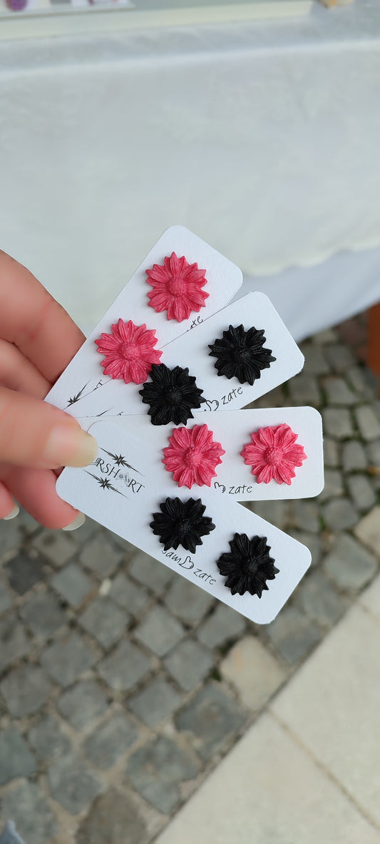 A hand is holding three white cards, each displaying a pair of handmade polymer clay earrings shaped like flowers. The earrings come in two colors: pink and black. Each card has the brand name "UrshArt" and the word "zate" written on it. The background shows a cobblestone pavement and a white tablecloth.
This image shows a close-up of handmade polymer clay earrings, highlighting their intricate flower design and vibrant colors, making it relevant for showcasing the craftsmanship and unique products.