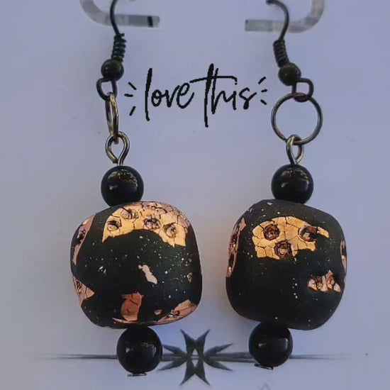 A pair of handmade earrings from UrshArt displayed on a white card. The earrings feature black beads with gold and copper flecks, creating a unique and artistic design. The card has the text "sam zate" at the top and "love this" in the middle, with the UrshArt logo at the bottom.
