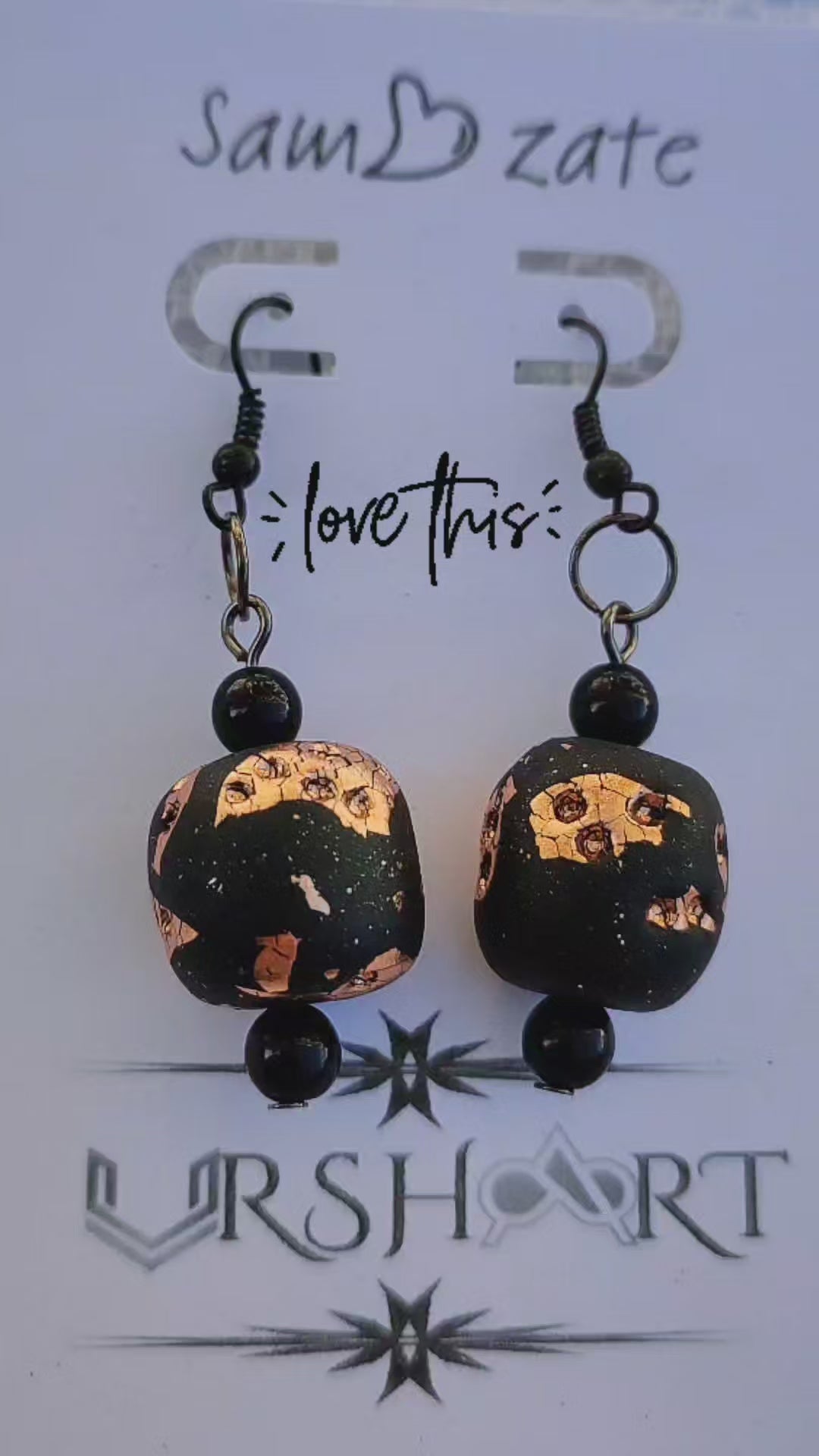 A pair of handmade earrings from UrshArt displayed on a white card. The earrings feature black beads with gold and copper flecks, creating a unique and artistic design. The card has the text "sam zate" at the top and "love this" in the middle, with the UrshArt logo at the bottom.
