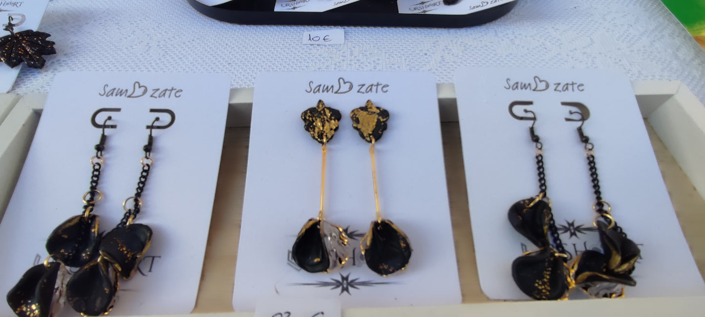 The image shows a display of handmade polymer clay earrings from a card for the UrshArt online store. There are three pairs of earrings on display, each attached to a white card with the brand quote "Sam zate" printed on it. The earrings feature intricate designs with black and gold accents. The pair on the left and right have black chains and dangling black and gold clay pieces, while the middle pair has a more elongated design with gold accents.