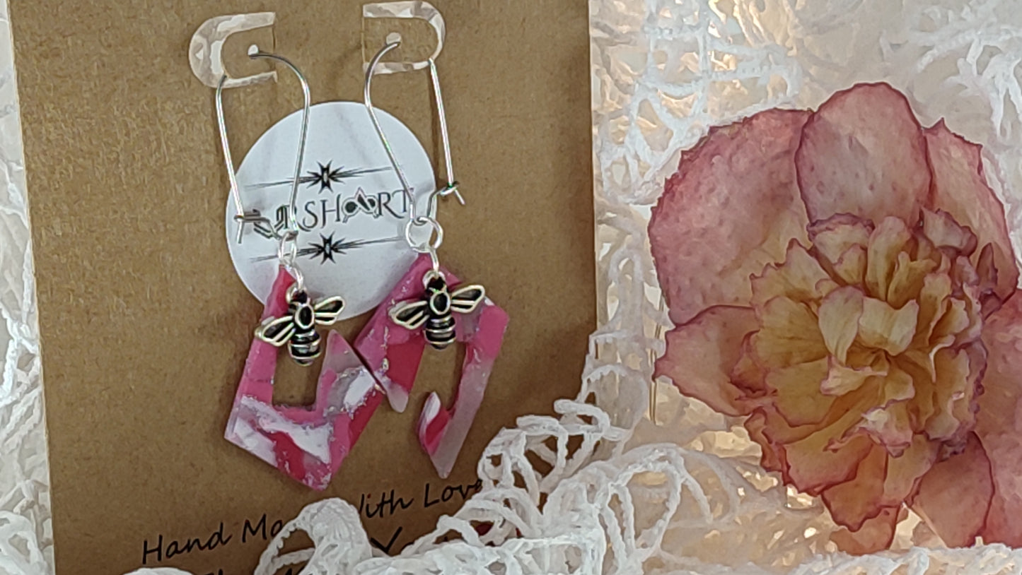 A pair of handmade earrings displayed on a brown card with the brand name "URSHART" printed on a circular white label. The earrings feature pink and white marbled geometric shapes with small bee charms attached. The card also has the text "Hand Made With Love" written on it. The background includes white decorative material and a pink and yellow flower.