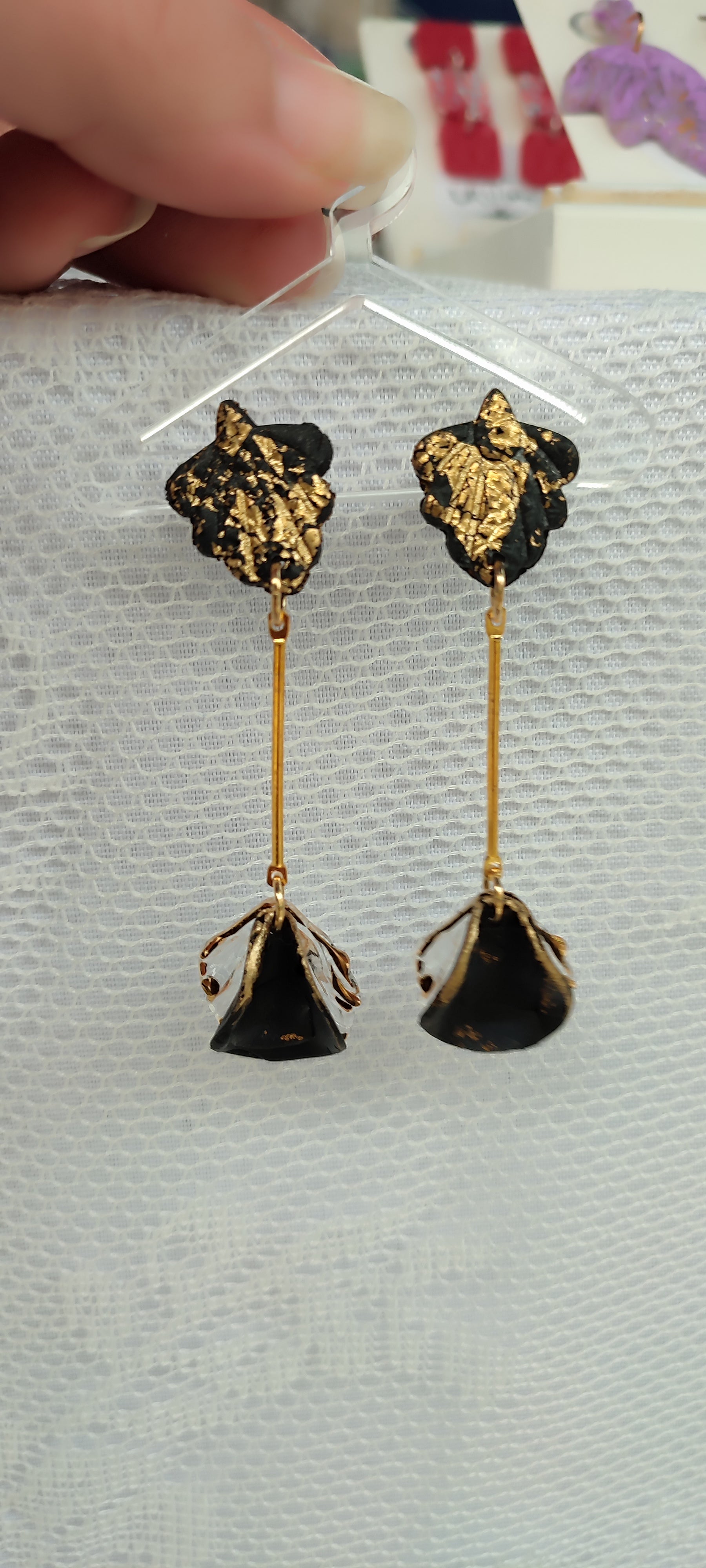 A pair of handmade polymer clay earrings displayed against a white textured background. The earrings feature a black and gold leaf design at the top, connected by a thin gold rod to a black and gold flower-shaped dangle at the bottom. The intricate detailing and combination of black and gold colors give the earrings an elegant and unique appearance.