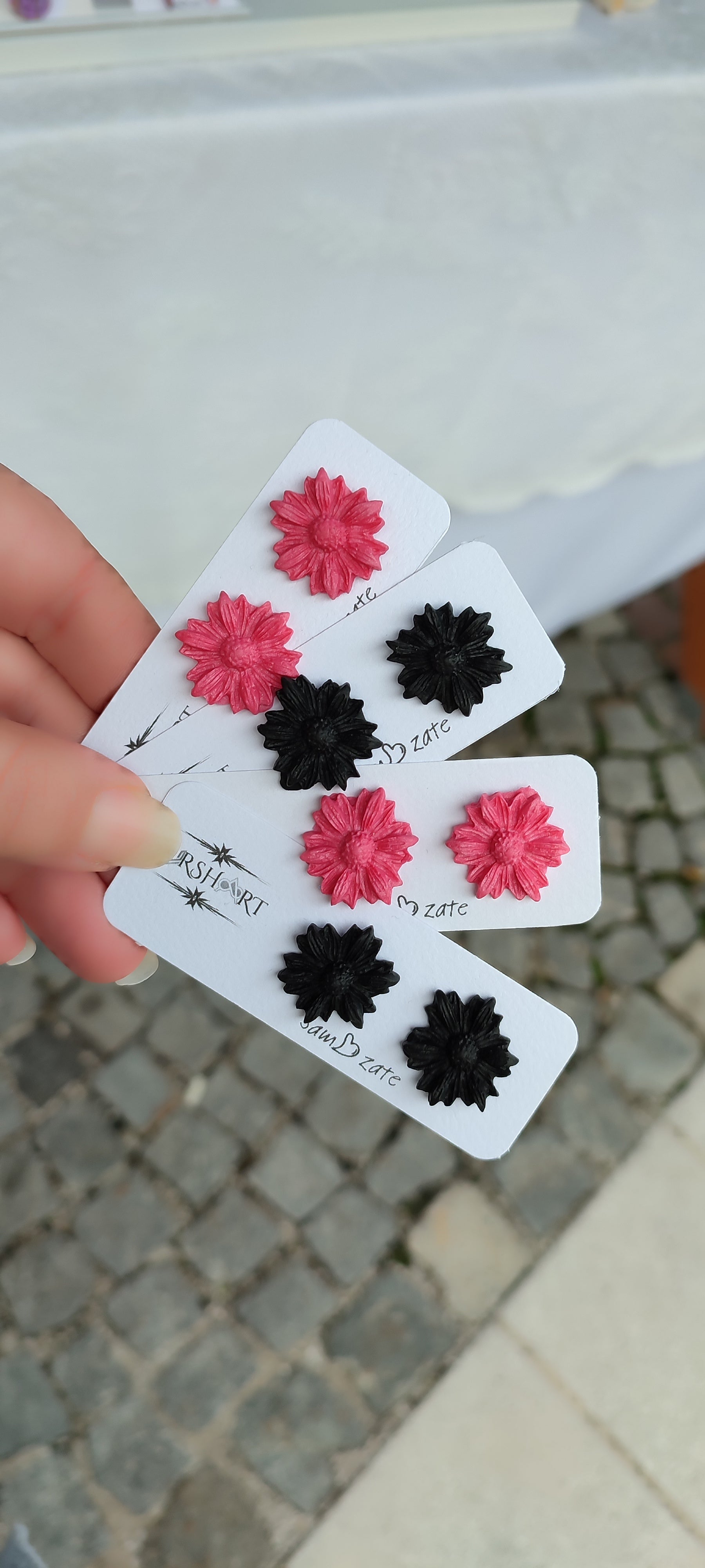 A hand is holding three white cards, each displaying a pair of handmade polymer clay earrings shaped like flowers. The earrings come in two colors: pink and black. Each card has the brand name "UrshArt" and the word "zate" written on it. The background shows a cobblestone pavement and a white tablecloth.
This image shows a close-up of handmade polymer clay earrings, highlighting their intricate flower design and vibrant colors, making it relevant for showcasing the craftsmanship and unique products.