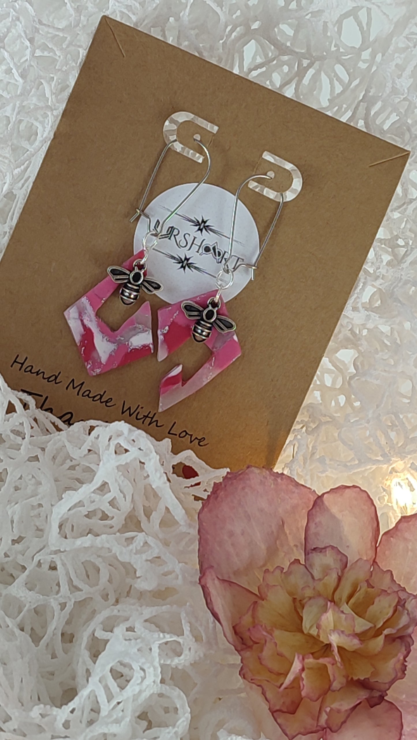 A pair of handmade earrings displayed on a brown card with the brand name "URSHART" printed on a circular white label. The earrings feature pink and white marbled geometric shapes with small bee charms attached. The card also has the text "Hand Made With Love" written on it. The background includes white decorative material and a pink and yellow flower.